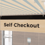 Self-checkout sign with black text and a black stripe on a wood grain background. Double sided from Shop2it Retail.